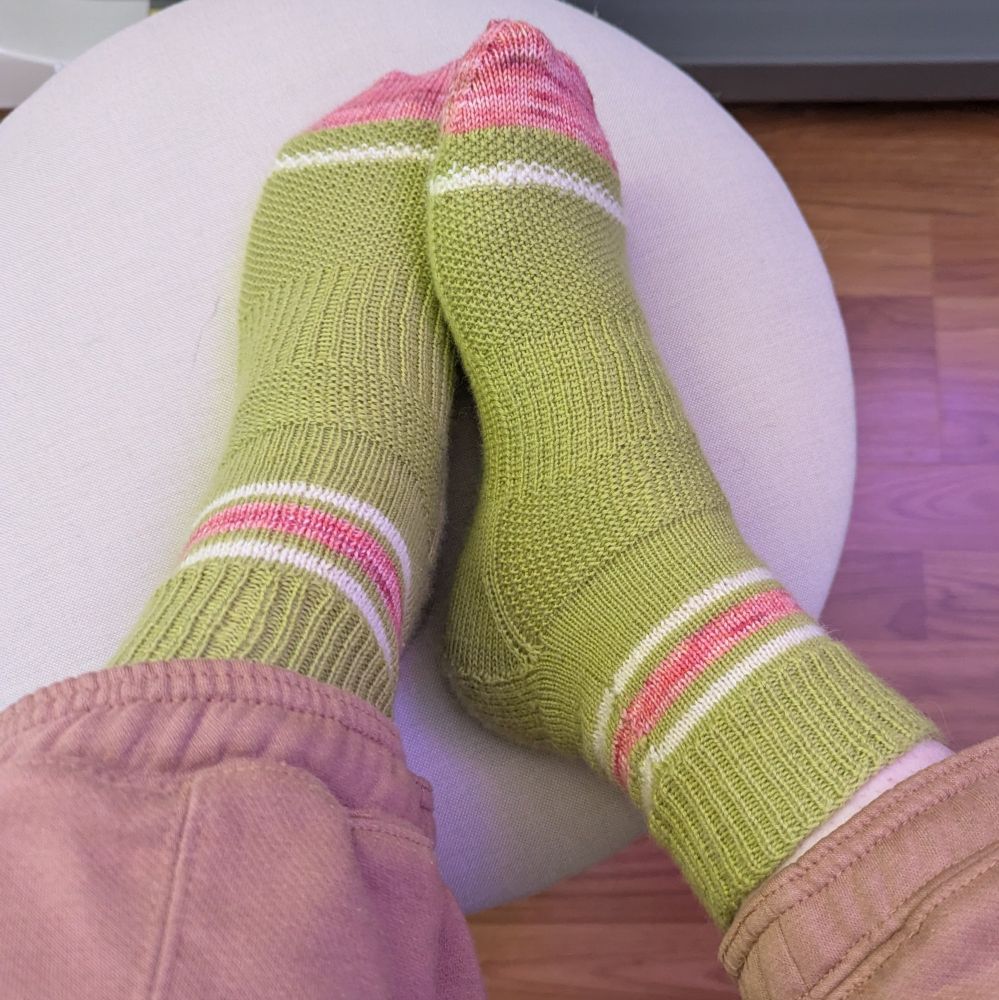 Photo of 2 brightly colored socks from top and side view on a woman's feet. They are avocado green with thin white stripes at the top with a thicker tonal reddish pink tone stripe in the middle. At the toe there is a thin white stripe before going into a tonal pinkish red toe.