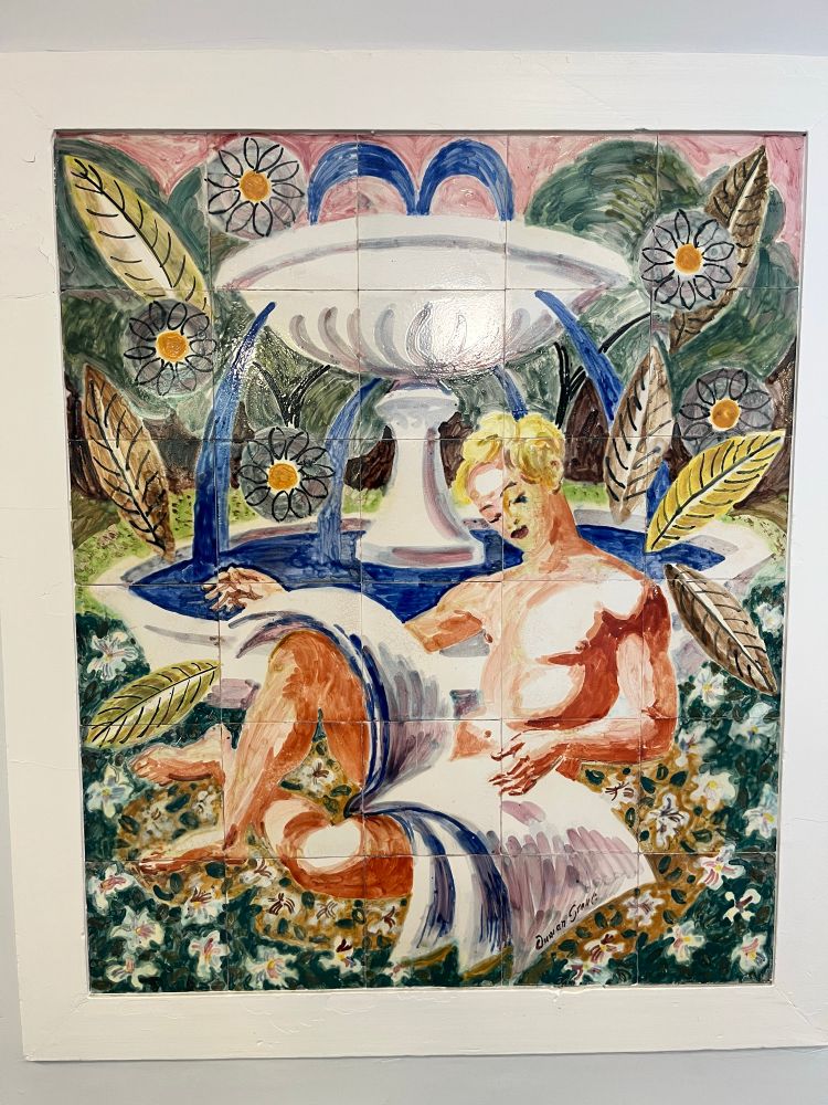 Painted ceramic tiles showing a naked young man with blond her reading the papers which cover the middle part of his body. He sits in front of a fountain on a meadow with flowers 