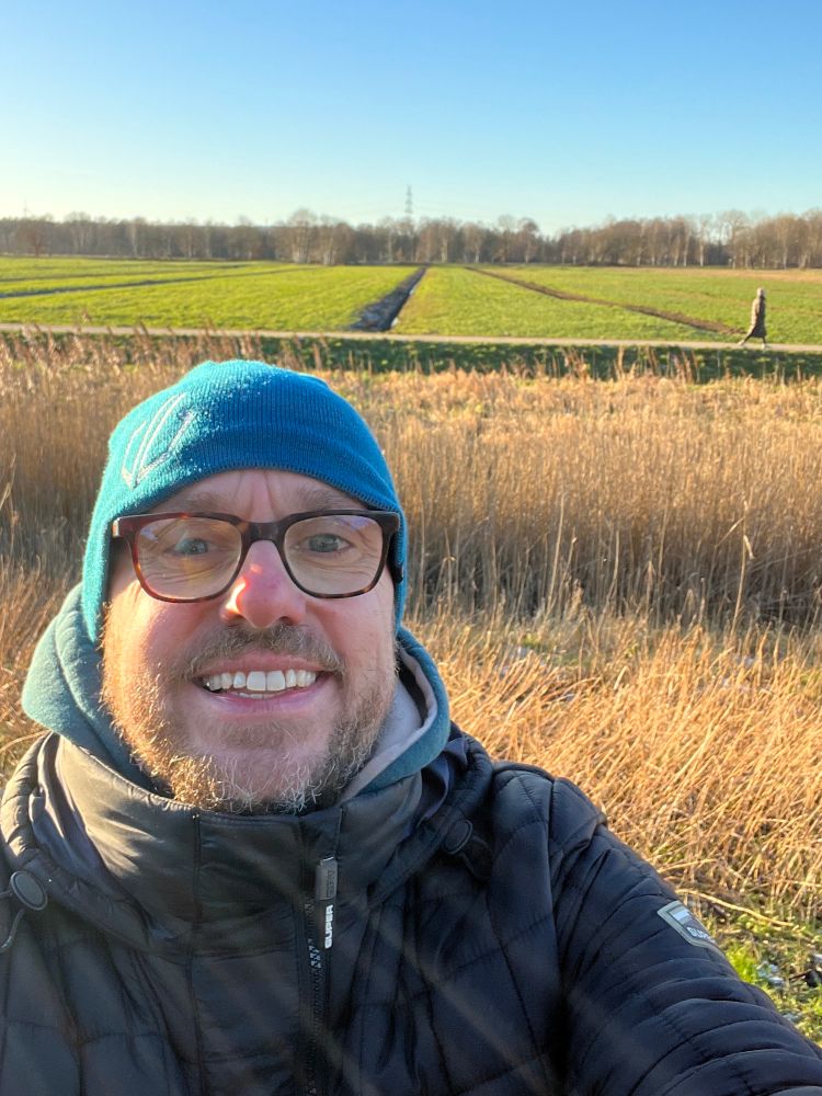 A selfie of middle aged man (me) with beard, glasses & a beanie with fields and a clear blue sky in the background.