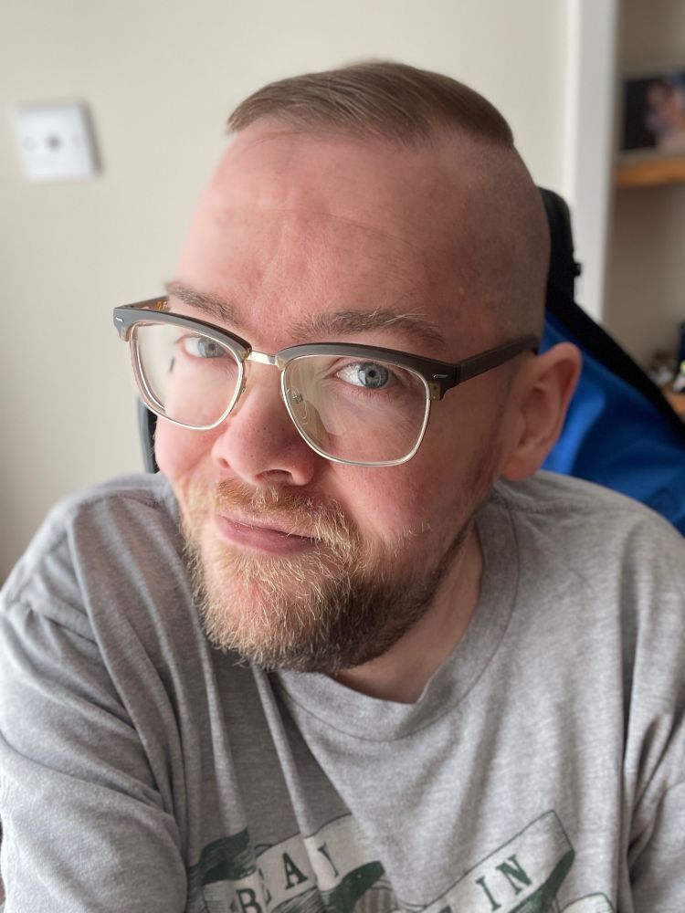 Selfie of Ally, a wheelchair user with a beard and glasses. They have a freshly shaved undercut. 