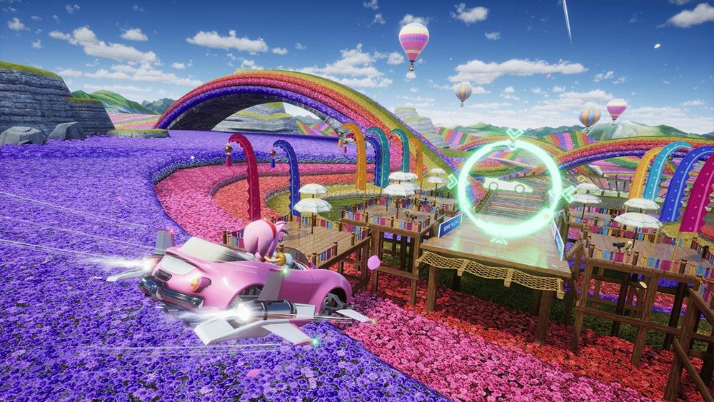 Amy Rose flying above a new location in her car, from the game Sonic Racing CrossWorlds. The location is covered in flowers of multiple colors, including purple, pink, and red. A bridge made of flowers can be seen in the distance. 