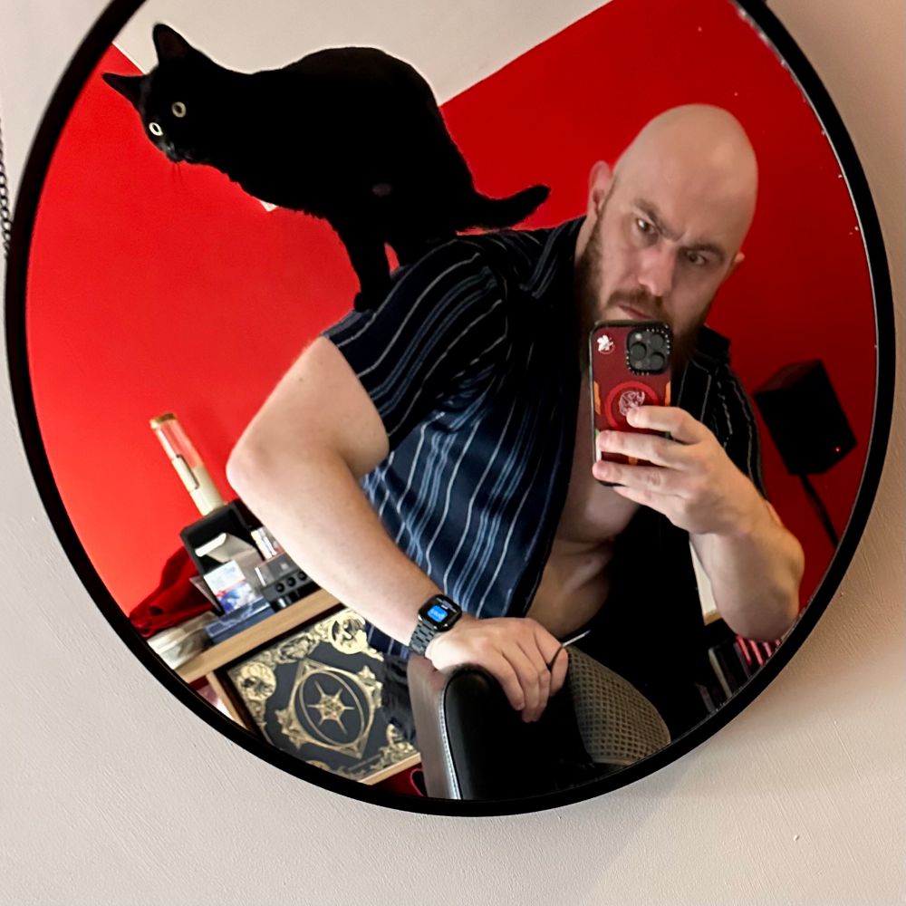 Kit stands in a mirror with his cat Dreadnought on his back.