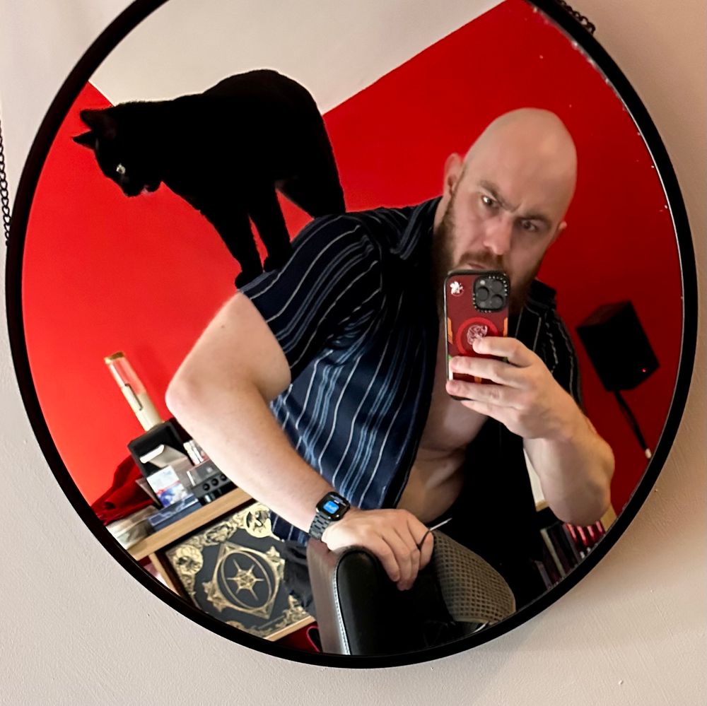Kit stands in a mirror with his cat Dreadnought on his back.