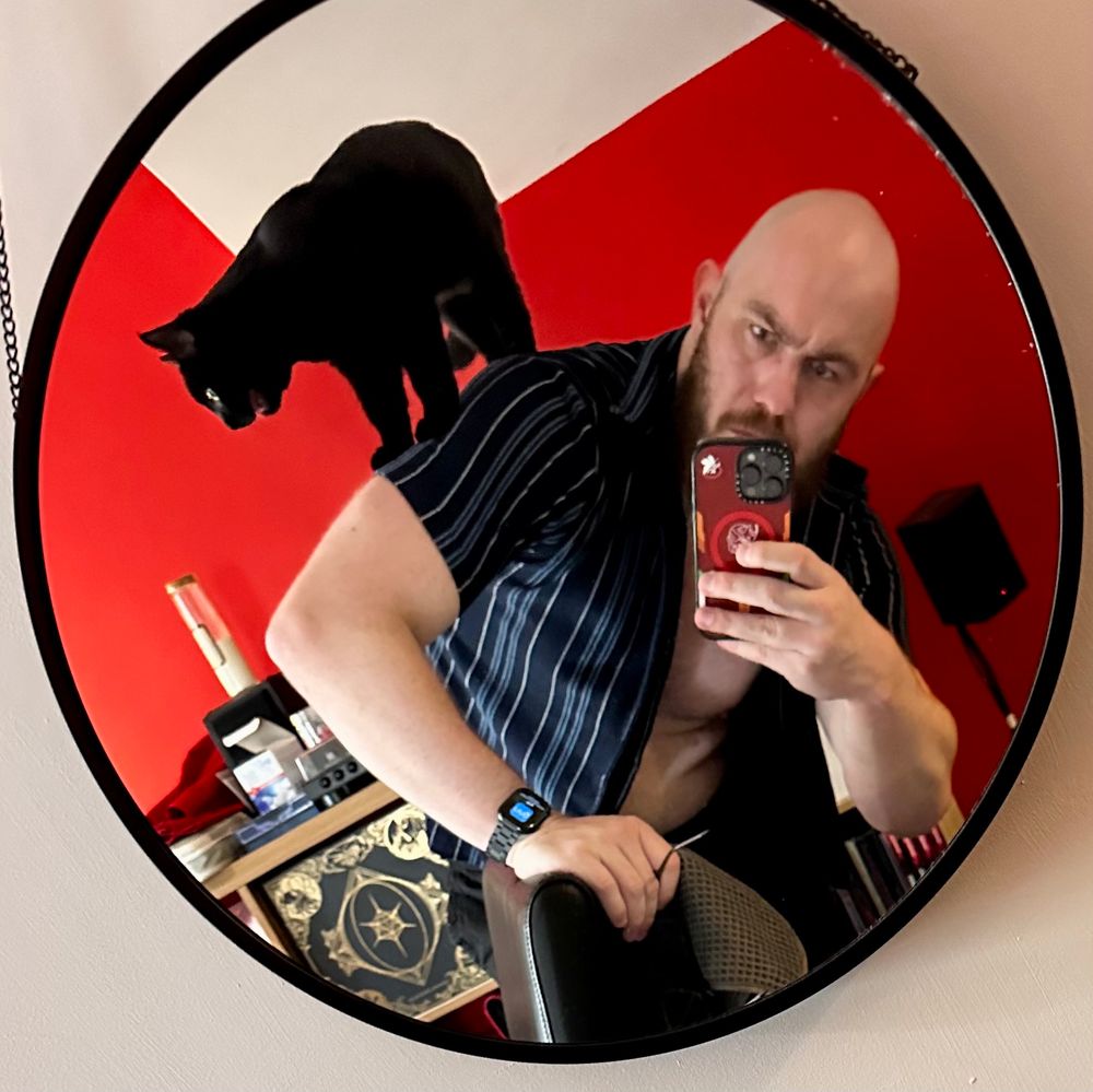 Kit stands in a mirror with his cat Dreadnought on his back.