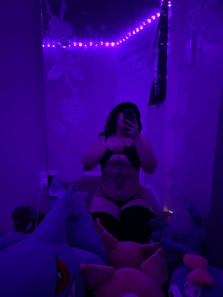 Dahlia posing in a mirror on her knees, wearing black thigh, highs, a black cami which is pulled up slightly to show her underboob, and a matching black thong. the lighting in the room is purple, and in front of her are various Kuromi stuffies and a large Gengar stuffie.