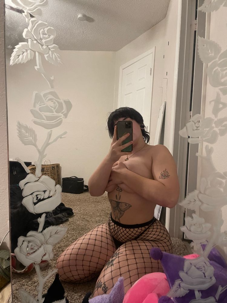 a mirror picture of Dahlia. her phone is covering her face but she is only wearing a black thong and fishnets. she is holding her tits with one hand as she sits in front of the mirror. the walls of the room are white and various Kuromi stuffies are set up in front of the mirror. 