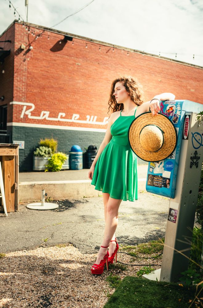 A woman with red hair and a green dress and red shoes stands in a bright sunlit scene, leaning against a blue payphone. There is a large brick building in the background. 