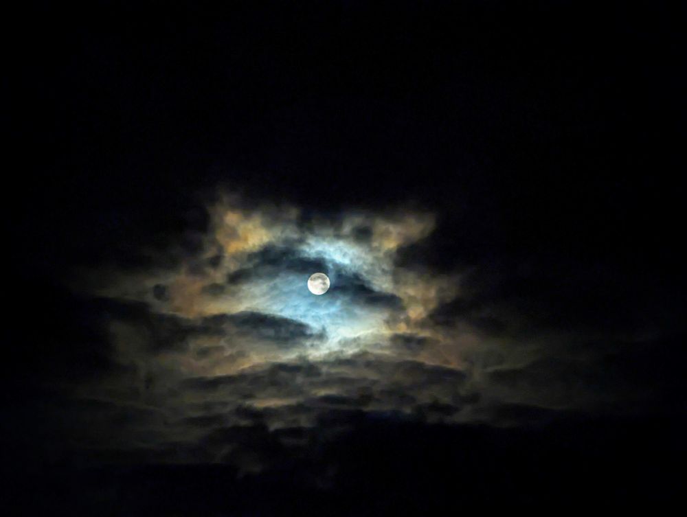 A wide view of a very bright full moon in the dark sky shining through quickly moving clouds.