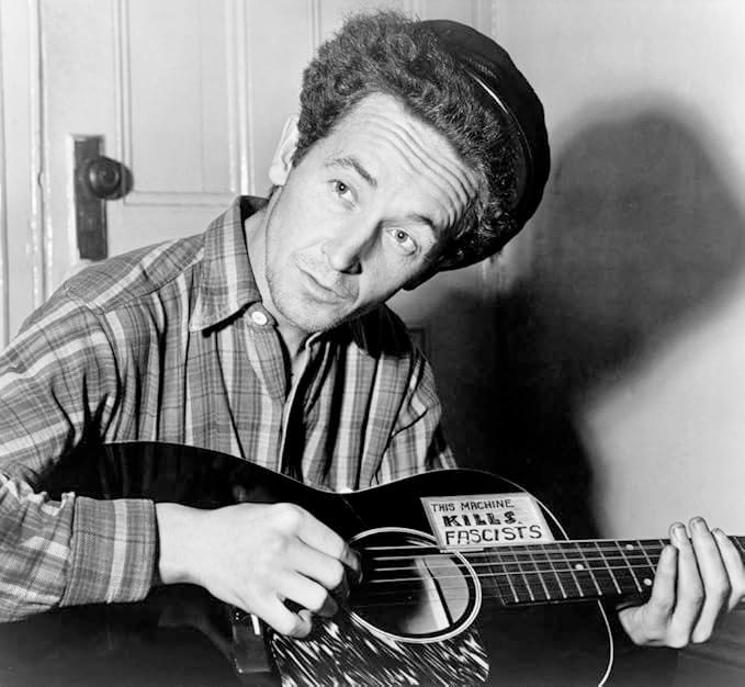 A black and white picture of Woody Guthrie in a striped shirt holding an acoustic guitar with a sticker on it that says "This machine kills fascists".