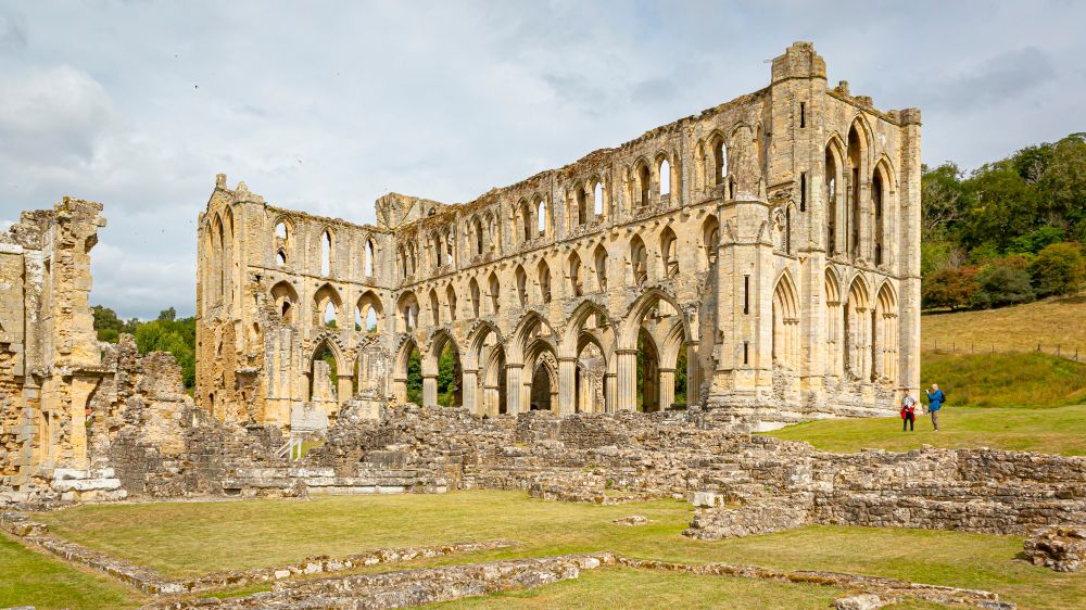 The exterior of a standing portion of a ruined abbey church constructed of light stone, featuring tall gothic arches and many pointed windows. Surrounding the church are the many ruined rubble walls of other abbey structures.