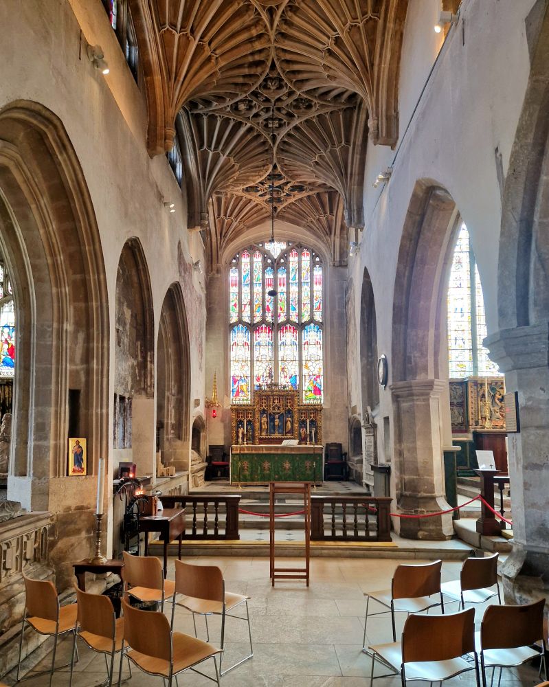 The interior of a narrow medieval church chapel. Either side is arcaded with tall stone arches set in white walls. At the far end is a tall window full of stained glass above an altar with a green cloth. The chapel ceiling has an unusual fan vault.