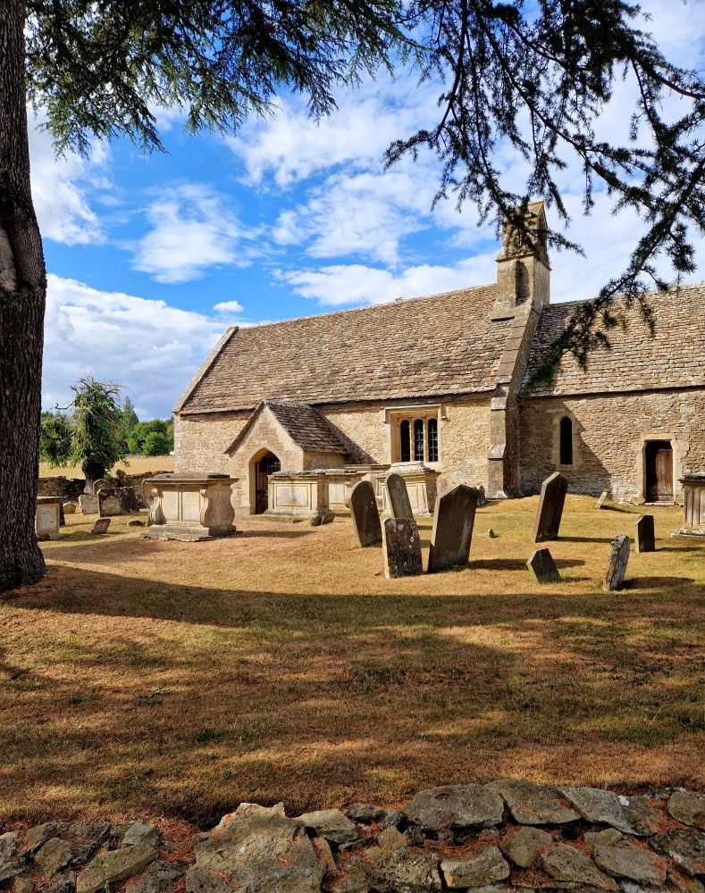 The exterior of the empty churchyard of a small rural medieval church built of light coloured stone, standing against a partly cloudy blue sky. The churchyard, dotted with many leaning headstones, stands in the shade is a tall cedar tree on the left of the frame. The grass is short and brown and dry.