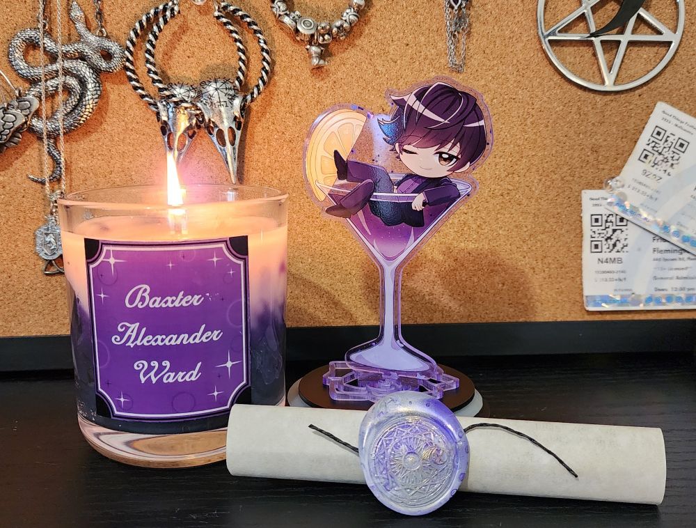 A candle made for the pansexual disasters that is Baxter Alexander Ward from OLBA with a standee of him sitting in a cocktail glass