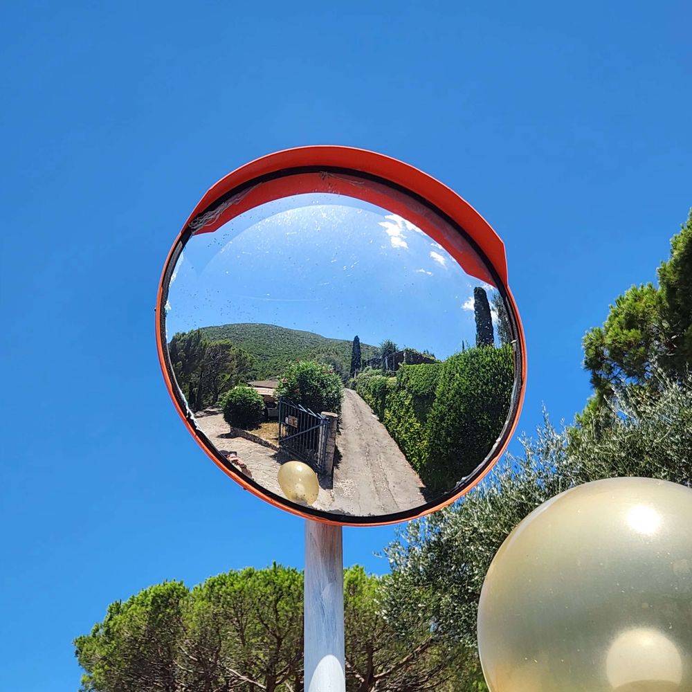 A reflective traffic mirror shows a sunny landscape with green trees and a winding path leading to a gate, under a clear blue sky. A golden sphere is visible in the foreground.
