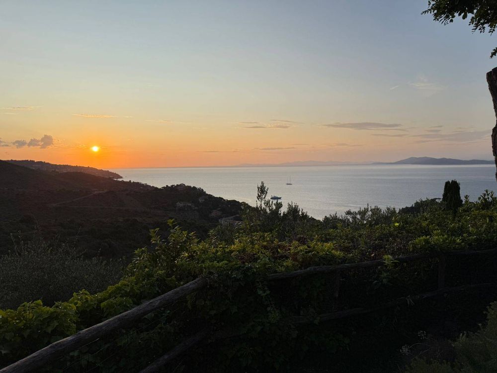 A serene landscape featuring a vibrant sunset over the calm sea, with silhouettes of hills and trees in the foreground, and a small boat on the water.