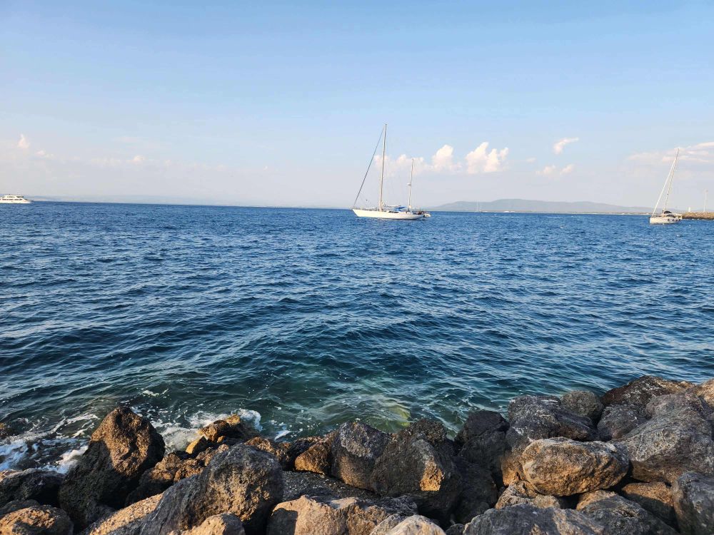 A serene view of the ocean with gentle waves lapping against rocky shores. Two sailboats are anchored in the distance under a clear blue sky with a few clouds.