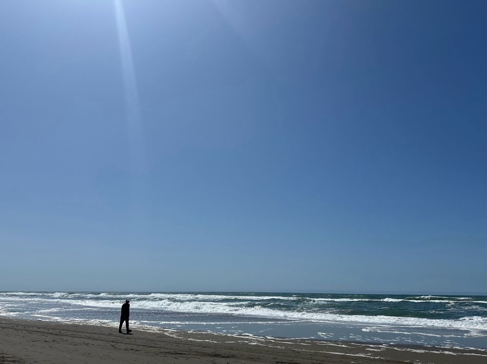 A walker along the beach as waves roll in and a blue sky dominates the view with sunlight streaming from the left 