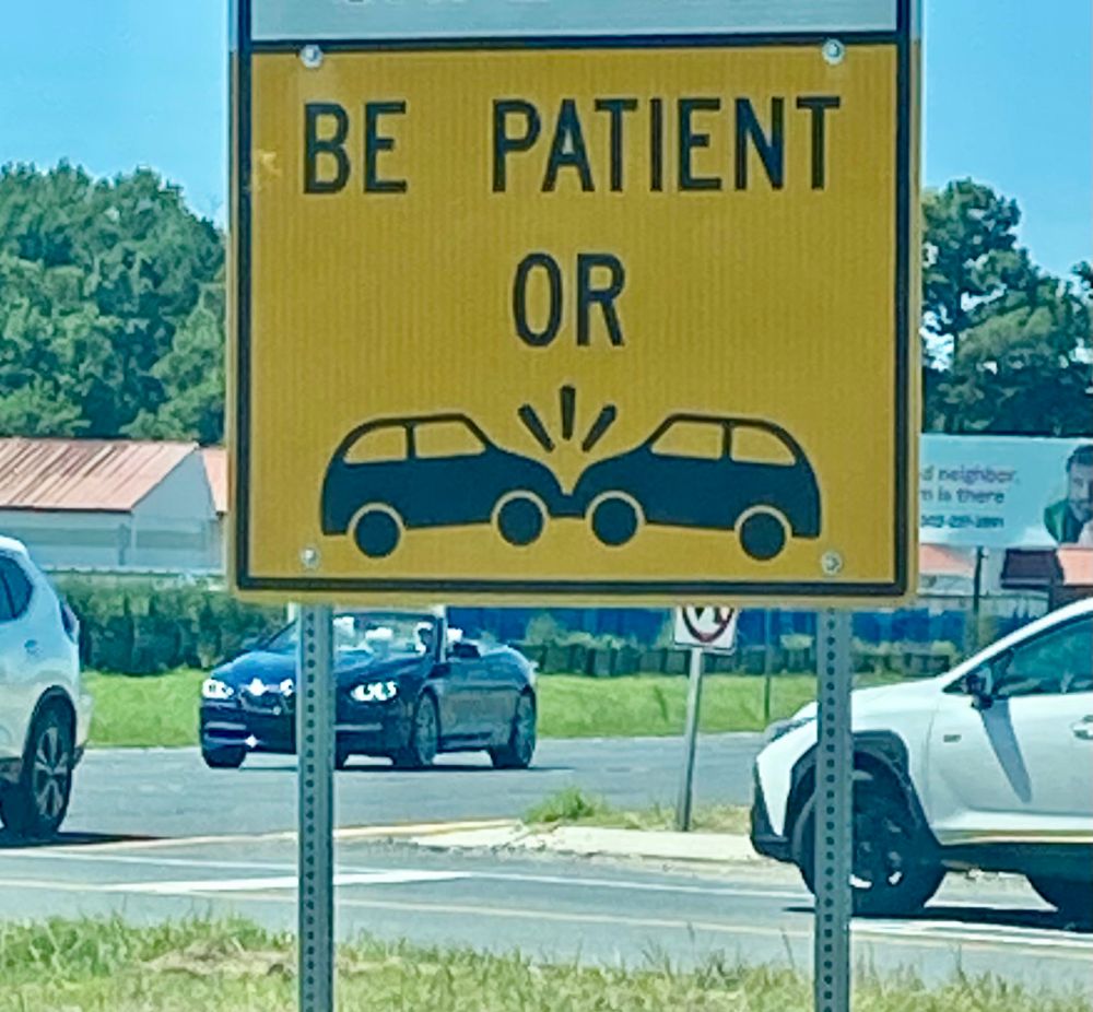 a road sign that says BE PATIENT OR and then shows two silhouettes of cars bumping into each other like they’re smooching 