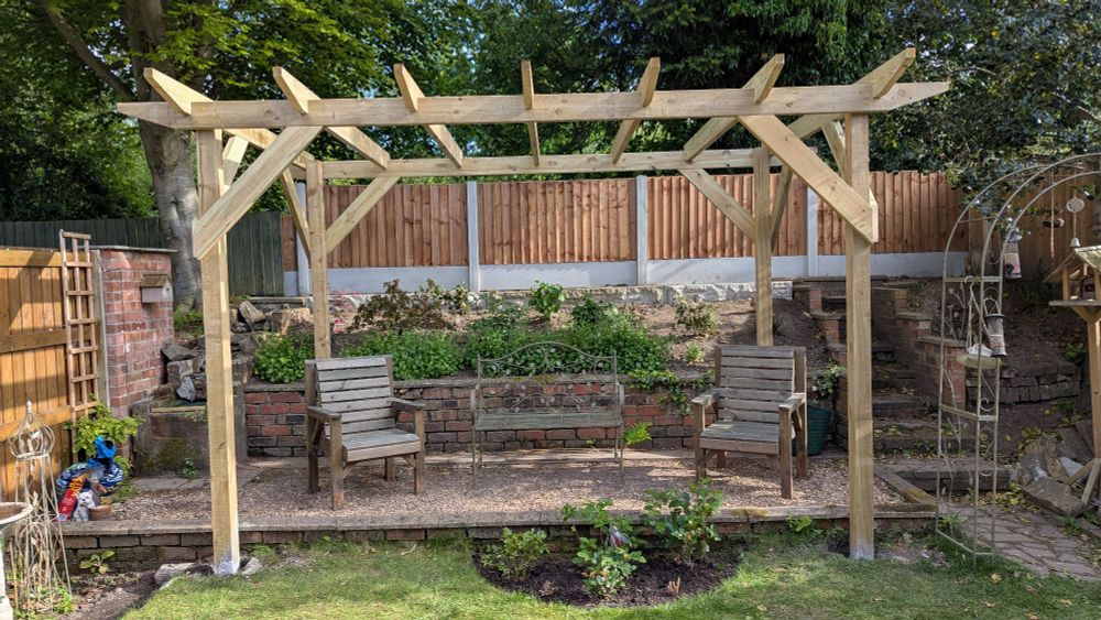 A wooden pergola in a garden, with a gravel floor underneath and green plants in a raised bed behind it. It has two wooden chairs and a metal bench underneath, and there is a small half-moon flowerbed in front of it with green plants in it.