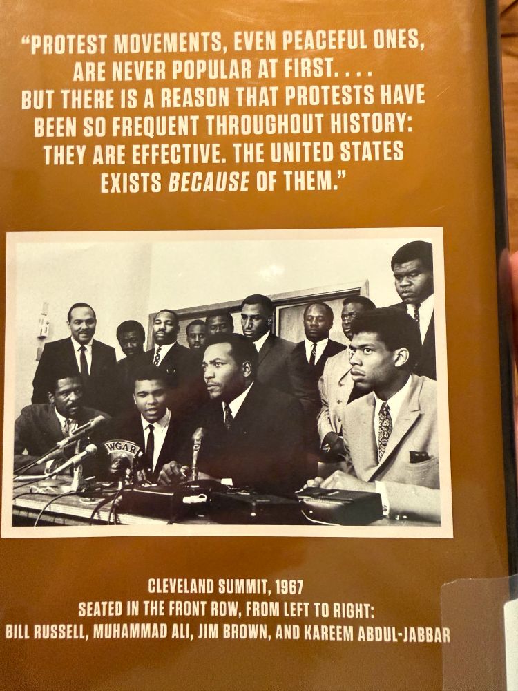 A framed photo and quote featuring the Cleveland Summit of 1967, a historic meeting of prominent Black athletes supporting Muhammad Ali’s refusal to be drafted into the Vietnam War. The top section displays a quote in white text on a brown background: “Protest movements, even peaceful ones, are never popular at first… But there is a reason that protests have been so frequent throughout history: They are effective. The United States exists because of them.” Below the quote is a black-and-white photograph showing several men in suits gathered around microphones at a press conference table. Seated in the front row from left to right are Bill Russell, Muhammad Ali, Jim Brown, and Kareem Abdul-Jabbar. The caption reads: “Cleveland Summit, 1967
