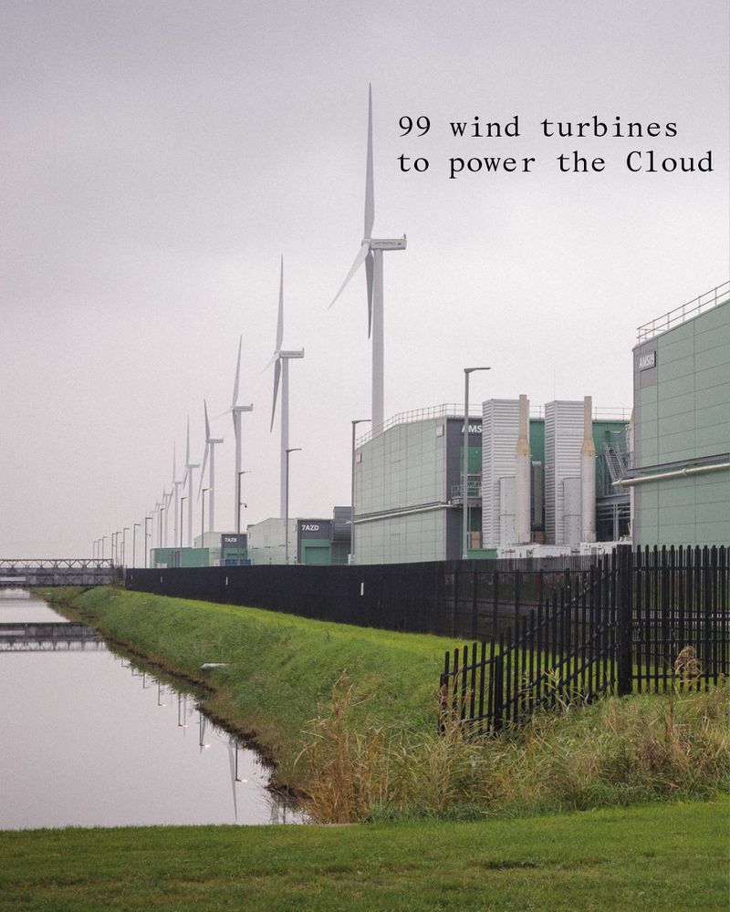 Long row of large wind turbines just behind a row of large data centres. 
