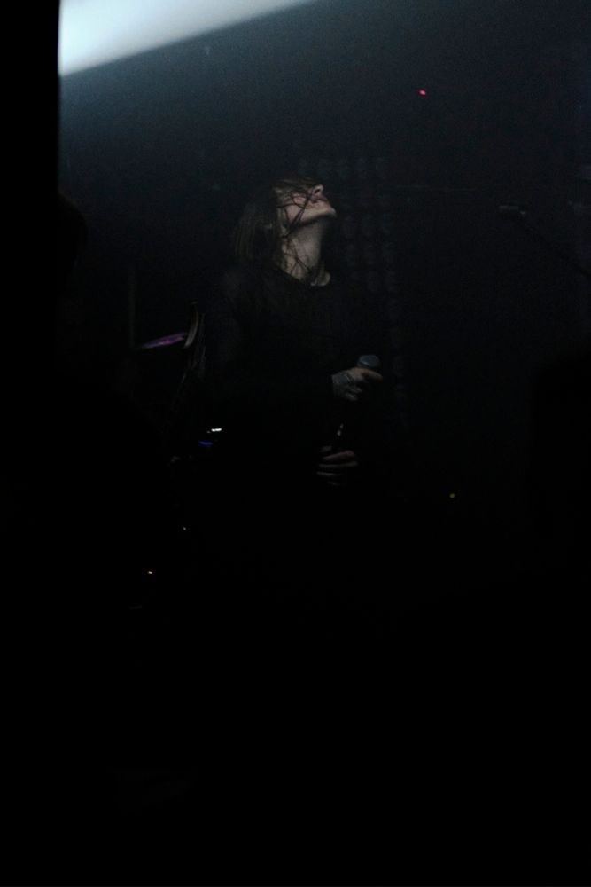 A person with long hair on a stage, the head tilted back, the sweaty hair clinging to their face. They clench a microphone, a bright light cuts through the surrounding darkness at the top