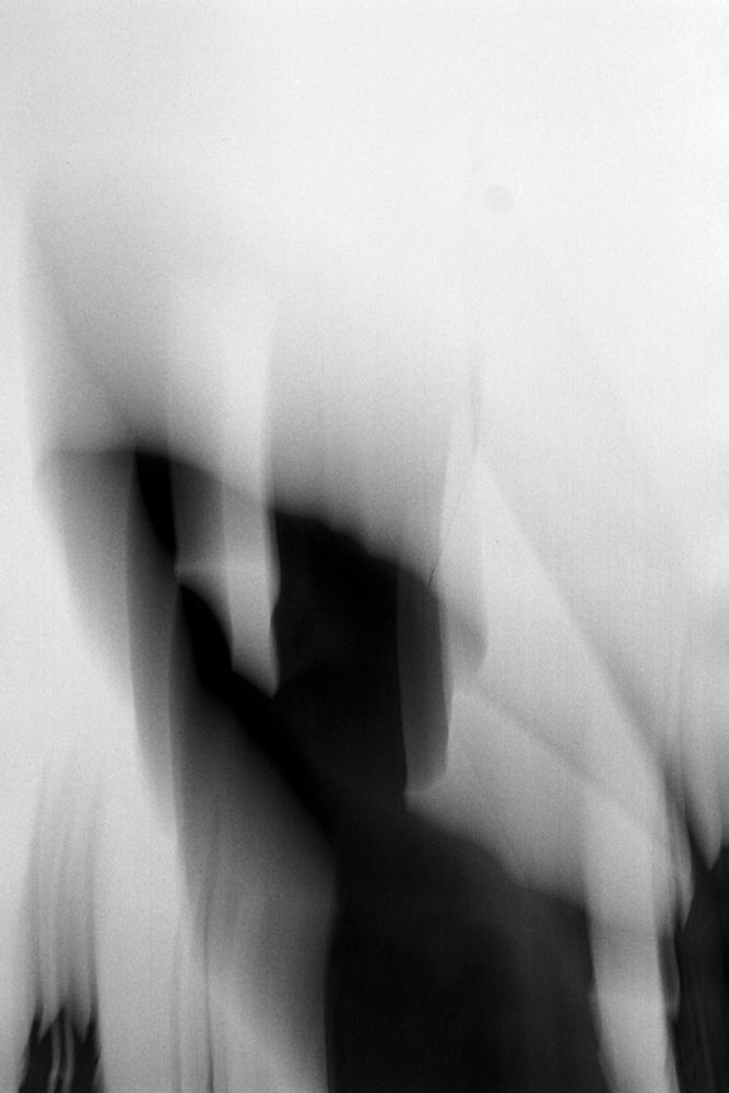 a blurry black and white photo of a mans silhouette wearing a helmet readying a bow shoot. parts of the silhouette is translucent a mist seems to emanate from it. A part of a building can be seen on the lower left, distorted and spikes of black mist