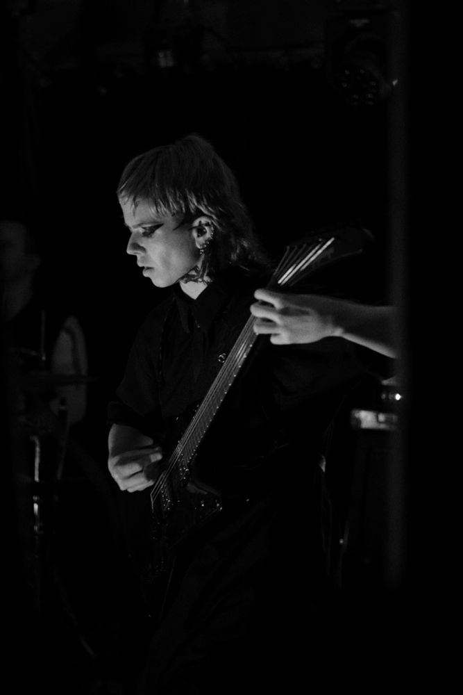 A blonde person with neck tattoos in black clothes playing electric guitar on a stage. They're very much in the flow
