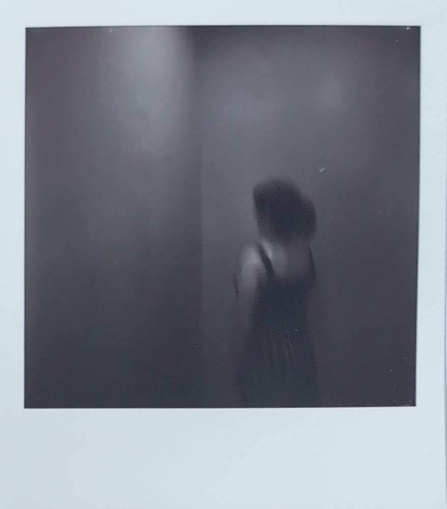 Ablack and white Polaroid of a person with a ponytail facing a corner, the person is blurry, the surroundings seem not to be