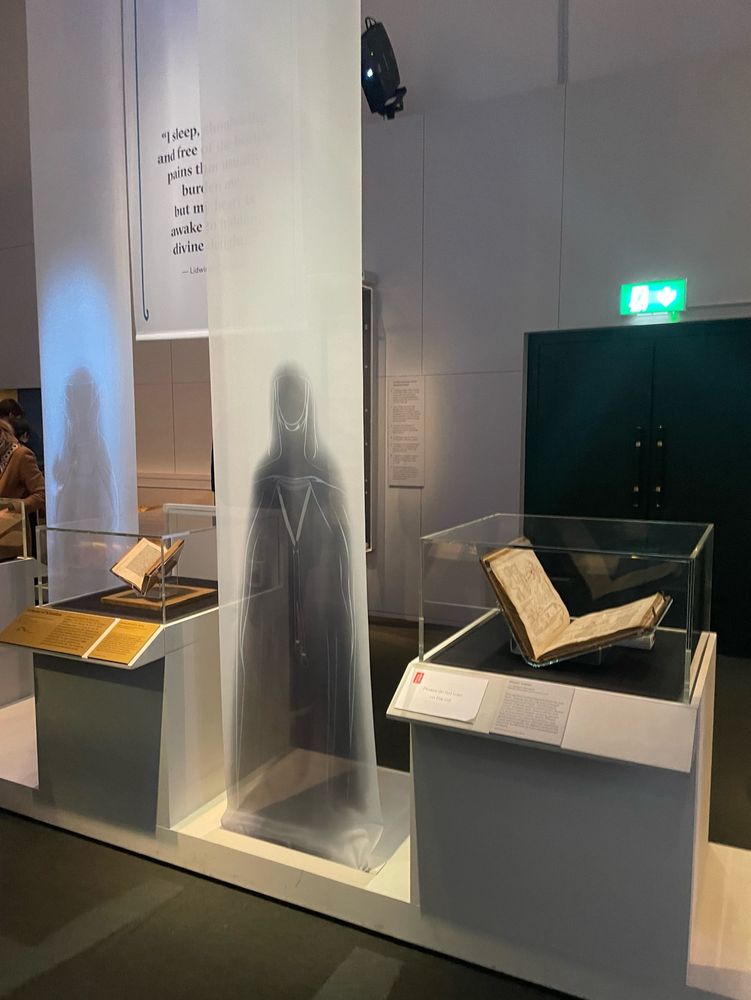 Installation shot of the exhibition at the British library, featuring two medical manuscripts. In between two cases is a ghostly figure, projected onto a sheer sheet of fabric hanging from the ceiling. 