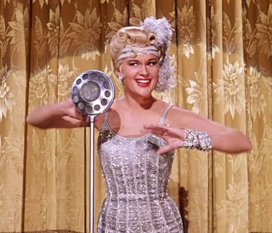 A cheerful woman dressed in a silver, spaghetti strap flapper dress and matching headband with a feather on her head of short, blond hair. She's standing in front of a gold theater curtain with a large, round, silver microphone on a stand in front of her.
As she smiles, her arms are up, bent at her elbows to each side, hands cupped in that old-fashioned flapper style.
Jean Hagen, born on this day in 1923 (d. 1977) as Lina Lamont in a scene from the film SINGIN' IN THE RAIN (1952)

Tags
Filmsky moviesky MOVIES films botd happy birthday Hollywood legends sexy beautiful actress MGM musical golden age comedy Oscar Academy Award nominee  