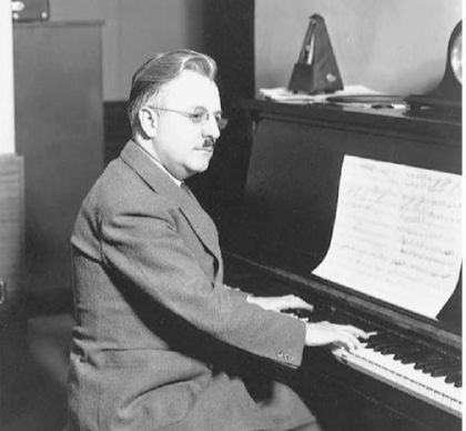 A black and white photo of composer Scott Bradley seated at a piano in a suit, hands on the keyboard. 
