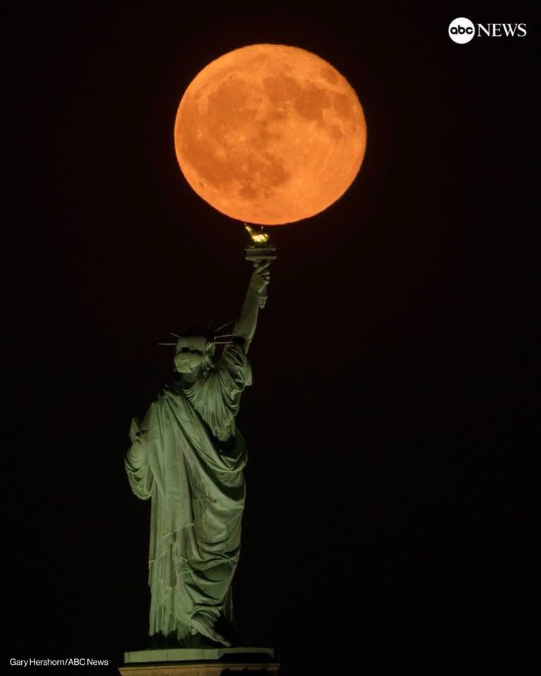 A photo of the Statue of Liberty taken from a low angle so it looks like the large orange full moon is sitting on top of the torch