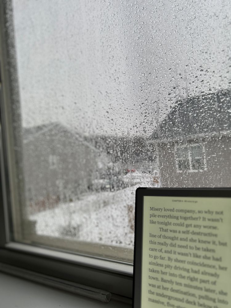 Picture of a window with raindrops on it and an e-reader in the corner. 
