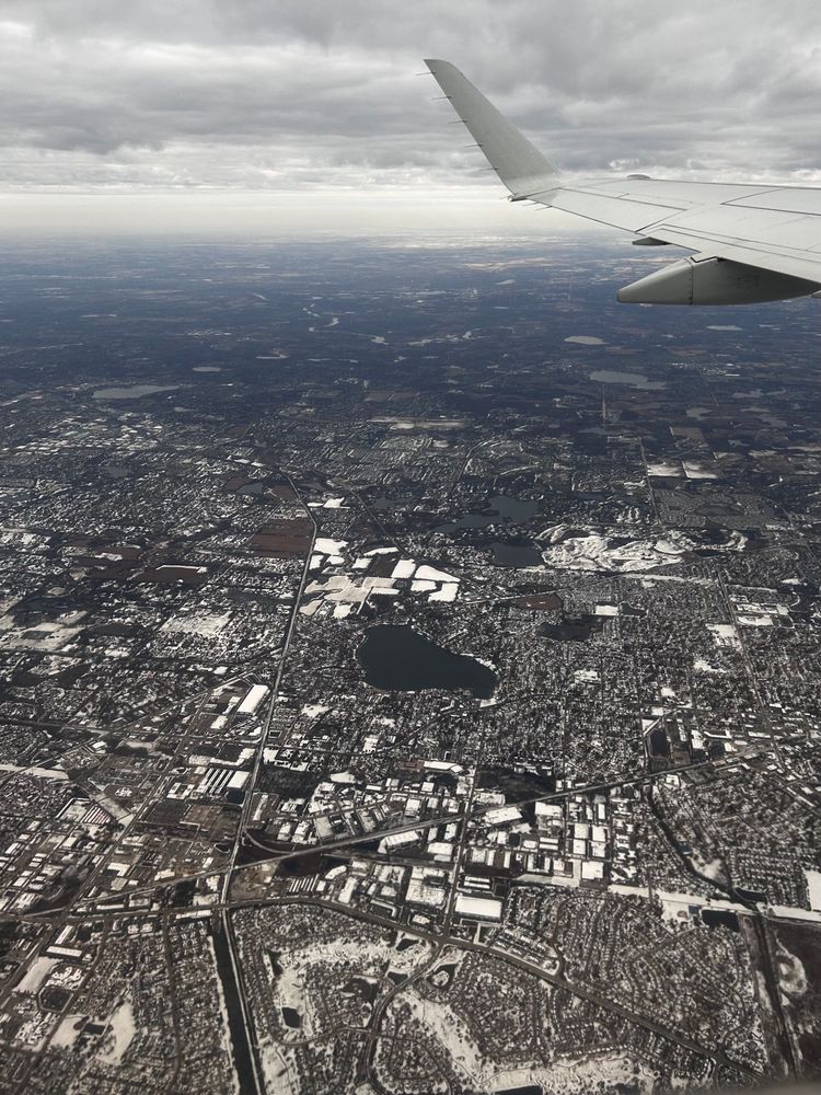 Suburban-ish development on low relief terrain with scattered small lakes. The snow cover gives roofs and grassy areas visual contrast. There's a portion of an airplane wing and low clouds. 