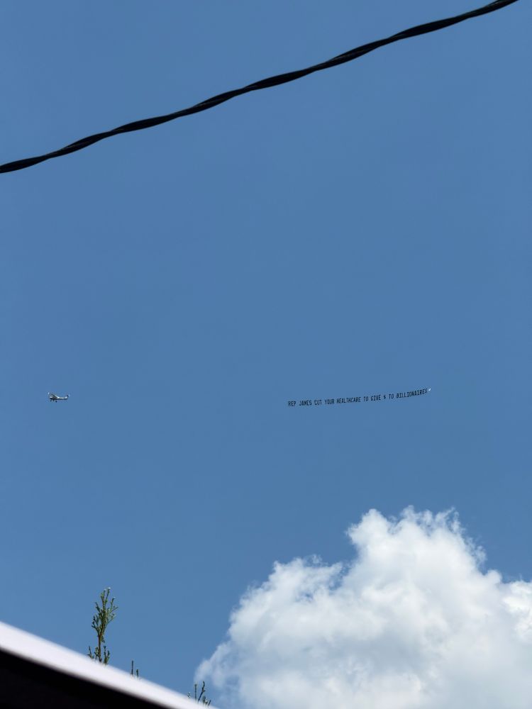 Airplane pulling a sign saying “Rep James cut your healthcare to give $ to billionaires”