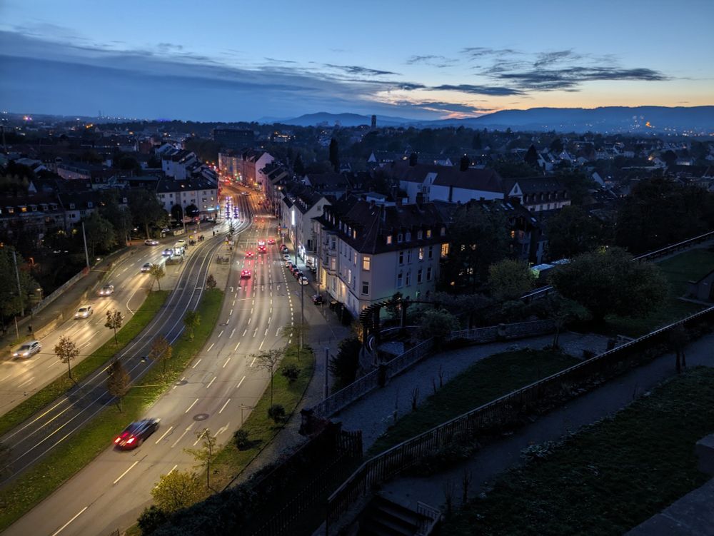 früher Abend, die Sonne ist schon untergegangen. Blick vom Weinberg in Kassel über die Weinbergterrassen, die im Schatten liegen, auf die sechsspurige Frankfurter Straße und auf viele Häuser. Man sieht neben der Straße Häuser, in denen hinter einigen Fenstern Licht an ist. Auf der Straße fahren einige Autos. Am Horizont sind Berge zu sehen