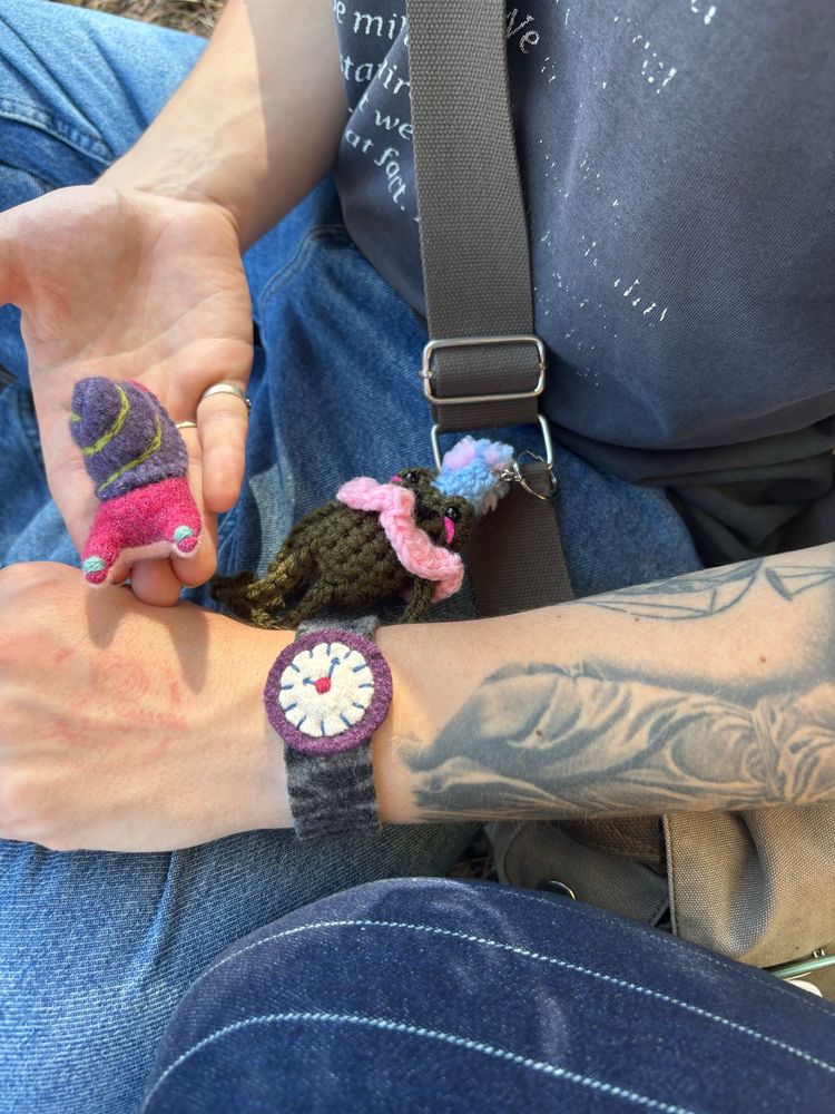 An arm with a felt watch, black strap with purple accents that reads 3:33, a small knit snail that is pink and purple, and a knit frog dressed as a jester attached to a bag.