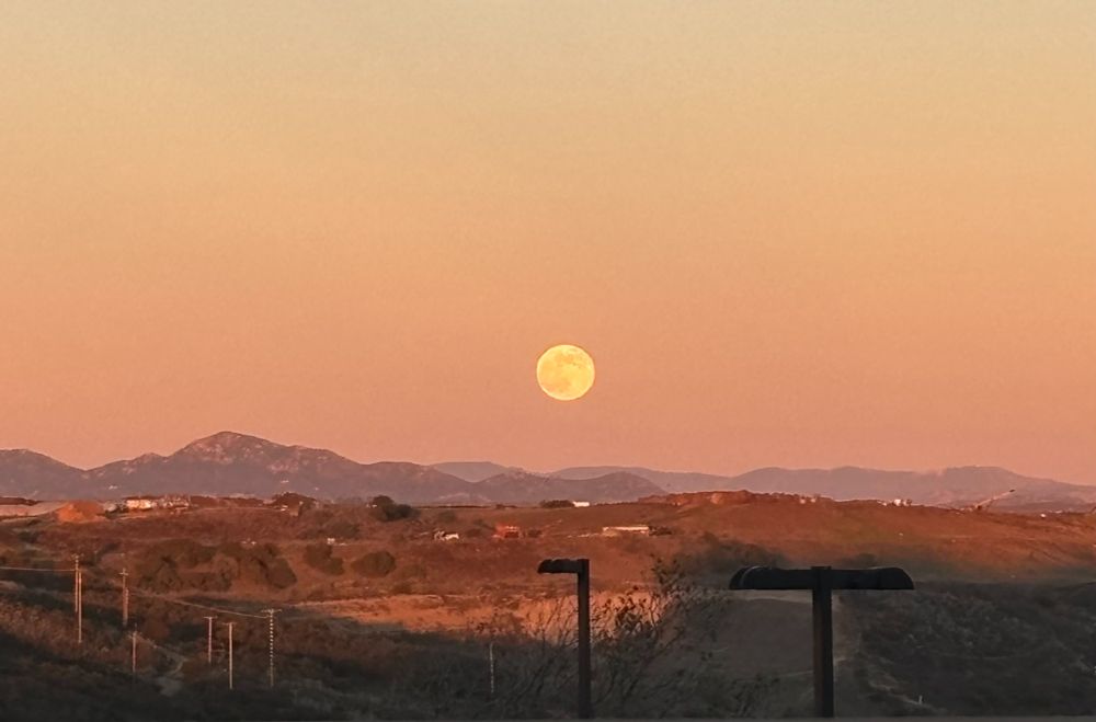 the moon rising over some mountains in a desert-y landscape
