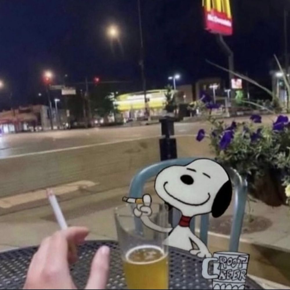 Image with the photographer holding a cigrette and a pint of beer. Across the table is Snoopy also holding a cigarette but drinking root beer. It’s night time on a city street and there’s a bright McDonald’s sign across the way 