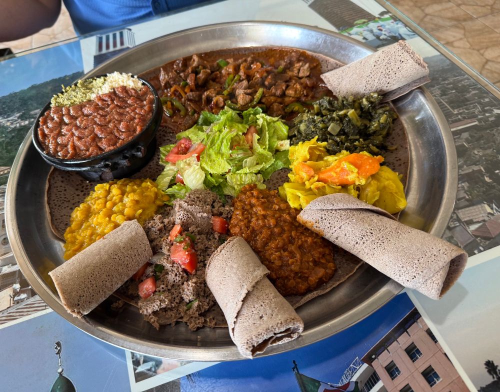 A plate of delicious Ethiopian food served on injera bread. There is Kitfo, red and yellow lentils, cabbage with carrots, collard greens, and awaze tibs. 