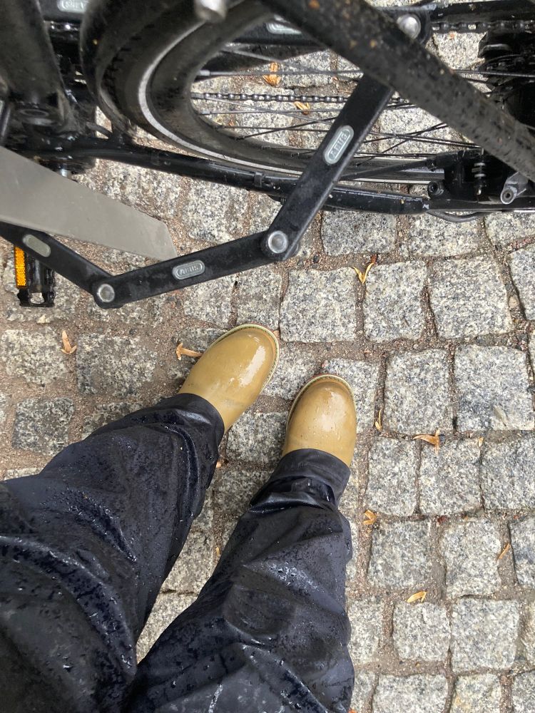 Hinterras eines Fahrrads, Beine in nasser Regenhose und nasse Gummistiefel.