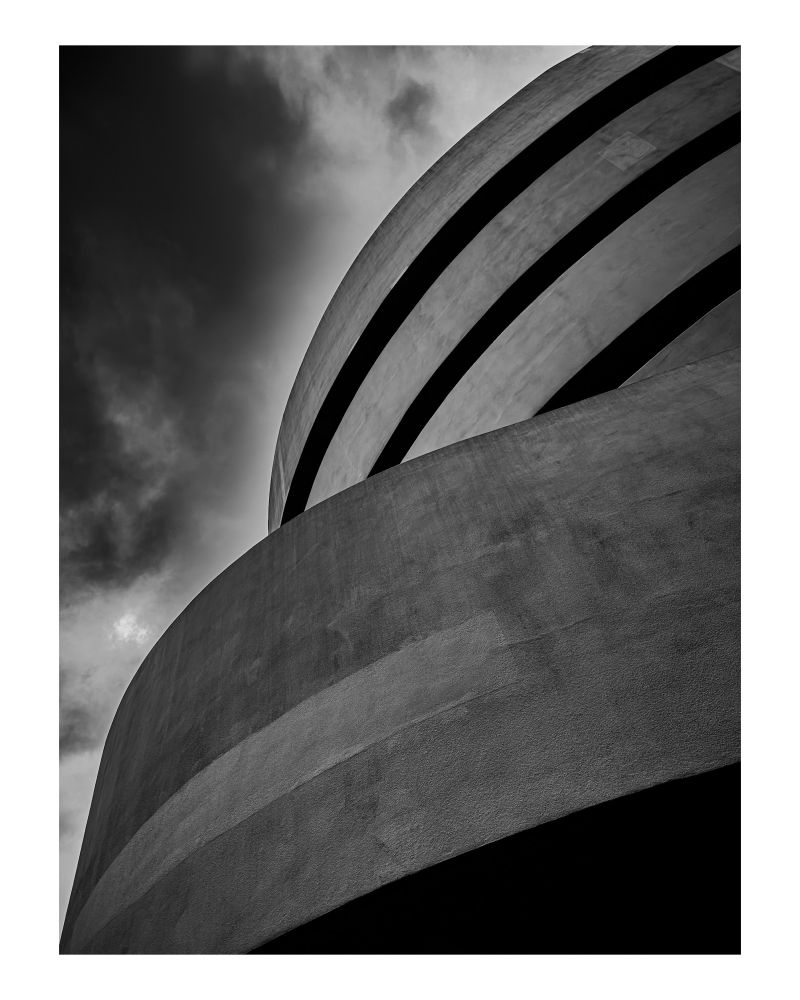 The curved exterior of the Guggenheim Museum in black and white, with dramatic clouds framing its smooth concrete texture.