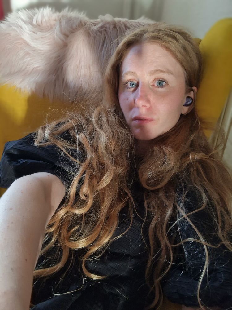 Kvinna med starkt solsken i fejset fotar sig själv i fåtölj. Har hon på sig en svart sopsäck? Svar nej det är en volangpuffelipuffklänning. Har hon druckit tequila å champagne? Svar ja 