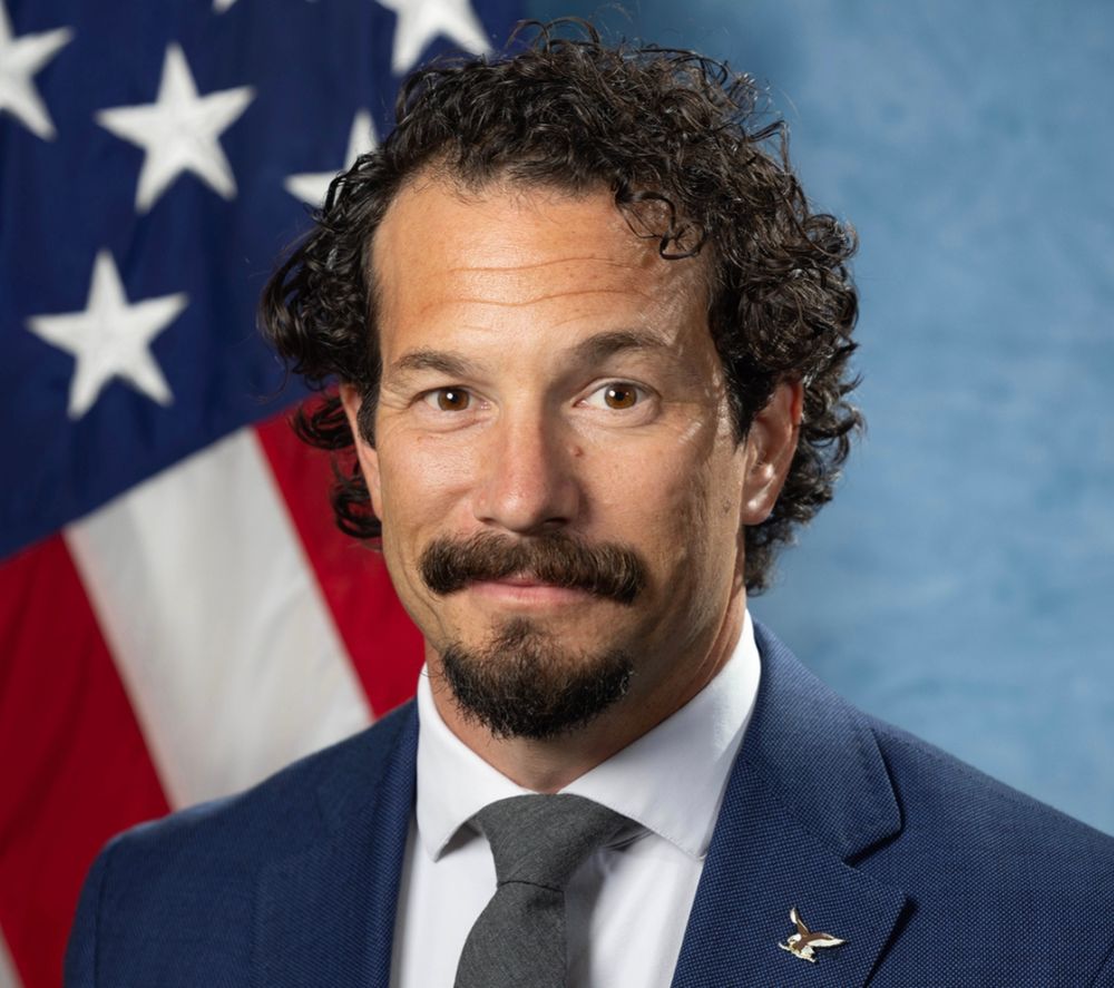Brian Driscoll, the acting director of the FBI, sporting curly hair, a kinda rakish jazz dirtbag moustache and beard combo, a faintly amused look in his eyes and a wry "whaddaya gonna do" smirk 