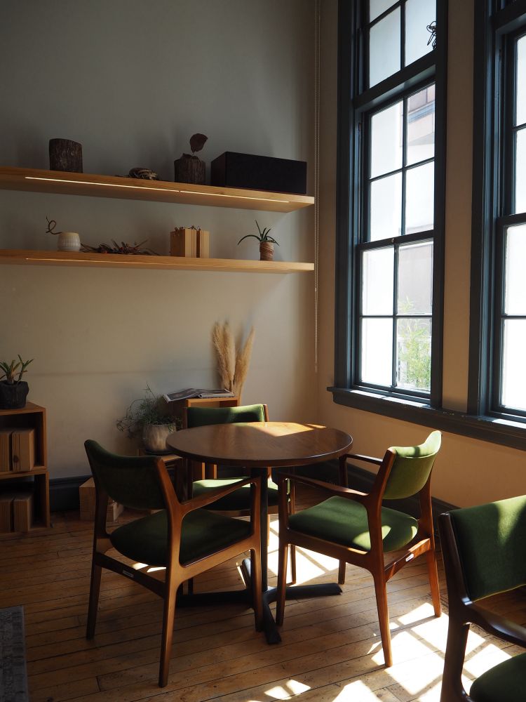 店内の写真です。中央に丸い木のテーブルがあり、囲むようにして椅子が三脚あります。椅子の背もたれや座面は緑色です。テーブルの後方右側には窓があります。