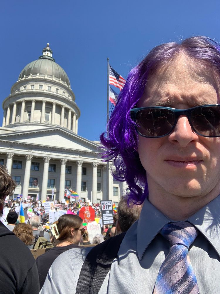 Toni at the 50501 protest wearing a gray shirt with a pink and blue tie. They have purple hair and sunglasses on. People are in the background holding protest signs. The Utah capitol building is also in the background.