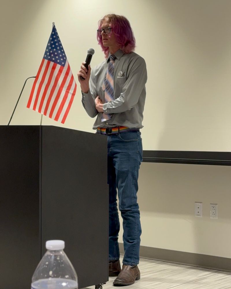 Tomkins standing behind a podium speaking into a microphone. They are wearing a gray shirt with a pink and blue tie and they have purple colored hair.