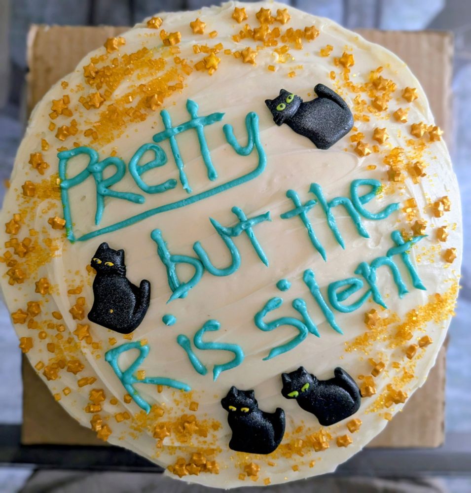 A layer cake with cream cheese frosting. In teal icing it says Pretty but the R is silent. There are 4 black cat royal icing decorations around the words and gold sprinkles with stars around the edges of the cake.