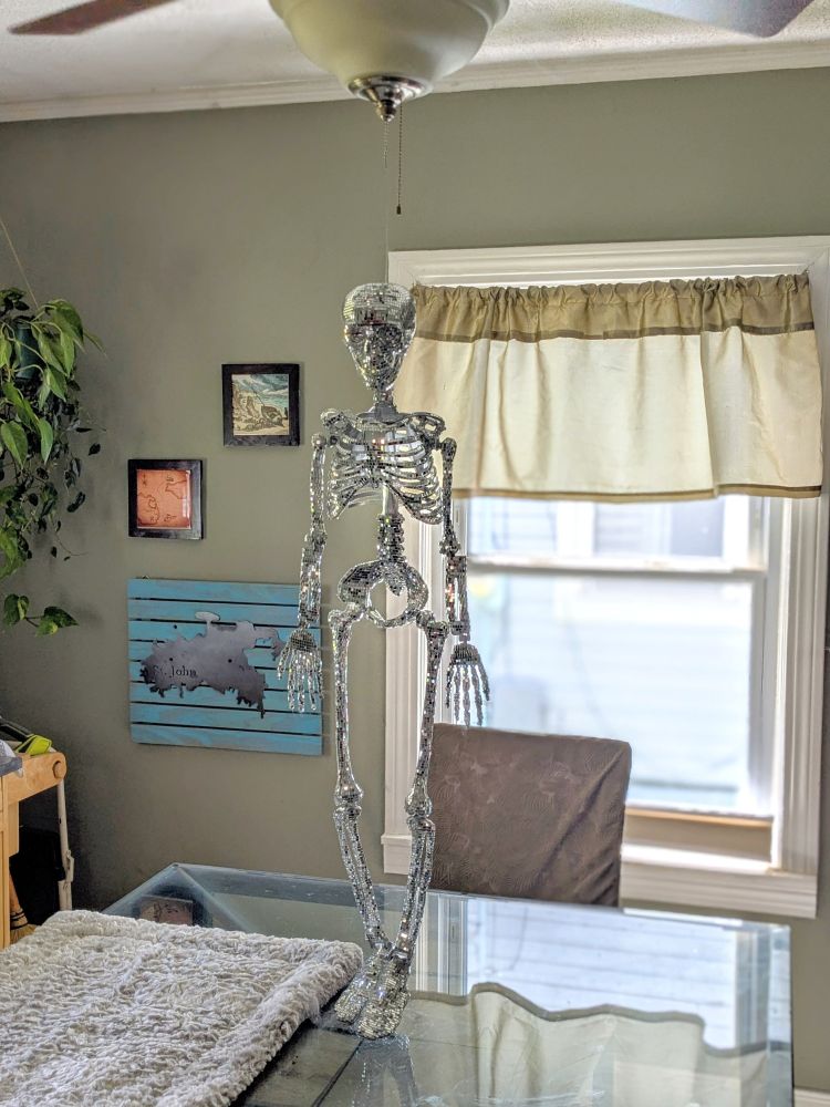 A 3 foot Skelton covered in little disco ball tiles hanging by clear cord from a ceiling fan so that her feet rest on the glass top of a dining room table. This picture is taken at a slight angle from the right.
